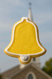 2.25 inch Bell Metal Cookie Cutter shaped yellow cookie with white icing against a church background.