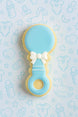 Cookie shaped like a baby rattle with a bow on a light blue background with baby-themed illustrations from a 4 inch Baby Rattle Metal Cookie Cutter