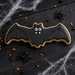A bat-shaped sugar cookie decorated with black icing and white icing details, placed on a dark wooden surface with Halloween props including spider decorations, orange and black sprinkles, and white cobweb from a 4.5 inch Flying Bat Metal Cookie Cutter