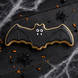A bat-shaped sugar cookie decorated with black icing and white icing details, placed on a dark wooden surface with Halloween props including spider decorations, orange and black sprinkles, and white cobweb from a 4.5 inch Flying Bat Metal Cookie Cutter