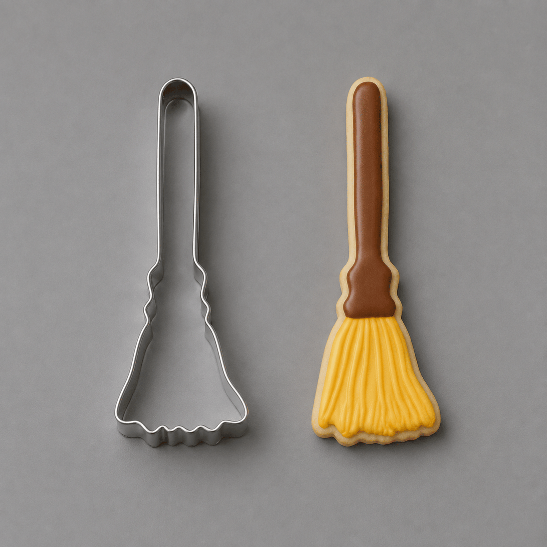 A metal witch broom cookie cutter shown next to a decorated sugar cookie shaped like a broom, on a solid gray background from a 5 inch Witch's Broom Metal Cookie Cutter
