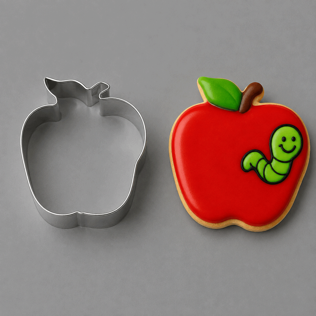 An apple-shaped metal cookie cutter shown next to a decorated apple-shaped sugar cookie with a red icing body, green leaf, brown stem, and a smiling green caterpillar from a 3 inch Apple Metal Cookie Cutter