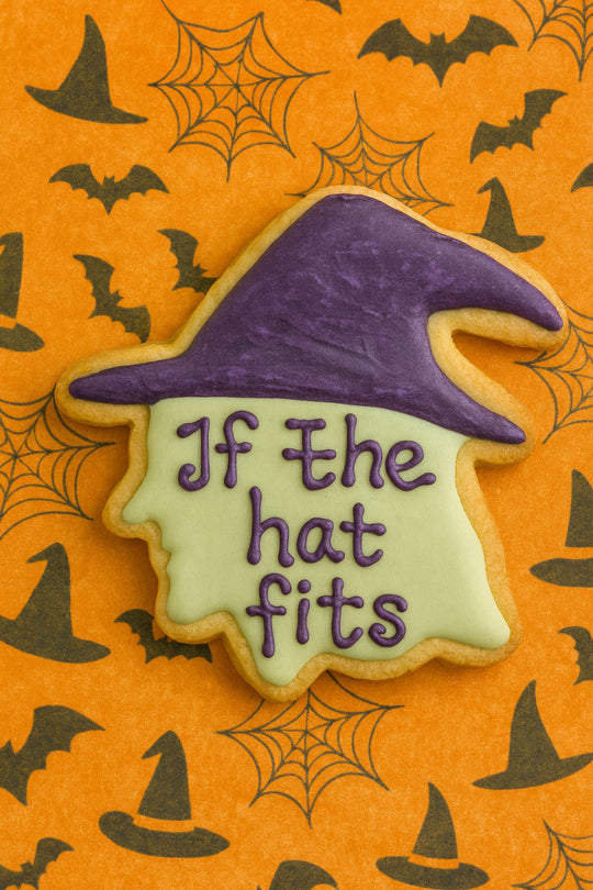 Halloween-themed witch head cookie with a purple witch hat and 'If the hat fits' text on an orange background with spider webs and bats.