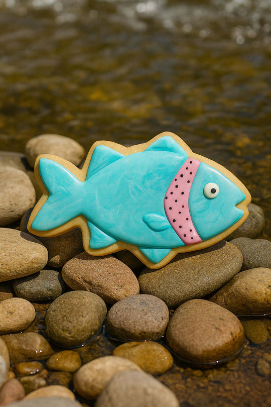 Fish-shaped cookie with blue icing and pink interior on pebbles near water from 5 inch Fish Metal Cookie Cutter