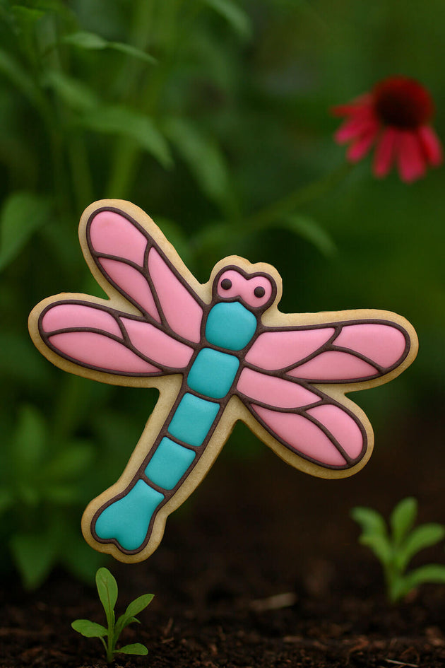 Colorful dragonfly-shaped object on a natural background with greenery and a red flower from a 3.5 inch Dragonfly Metal Cookie Cutter