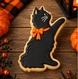 A black cat-shaped sugar cookie with an orange bow and green eye, decorated on a wooden surface with Halloween decorations including pumpkins and a lantern from a 4 inch Cat Metal Cookie Cutter