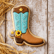 Decorative cookie shaped like a cowboy boot with blue foot and brown top, on a wooden surface with hay from a 3 inch Cowboy Boot Metal Cookie Cutter