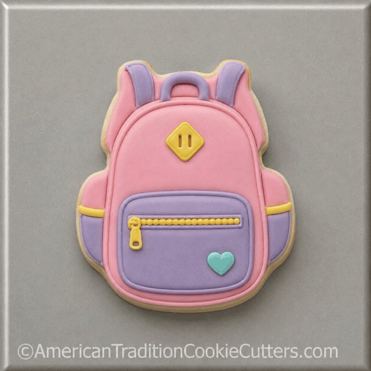 A pink and lavender sugar cookie cut in the shape of a backpack, decorated with yellow zipper details, a small light blue heart, and a yellow diamond emblem, set against a neutral gray background from a 3.5 inch School Backpack Metal Cookie Cutter