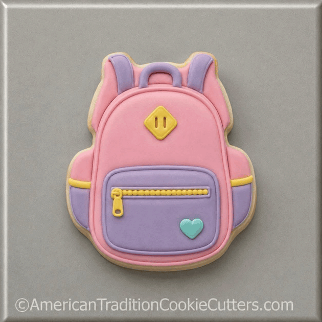 A pink and lavender sugar cookie cut in the shape of a backpack, decorated with yellow zipper details, a small light blue heart, and a yellow diamond emblem, set against a neutral gray background from a 3.5 inch School Backpack Metal Cookie Cutter