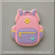 A pink and lavender sugar cookie cut in the shape of a backpack, decorated with yellow zipper details, a small light blue heart, and a yellow diamond emblem, set against a neutral gray background from a 3.5 inch School Backpack Metal Cookie Cutter