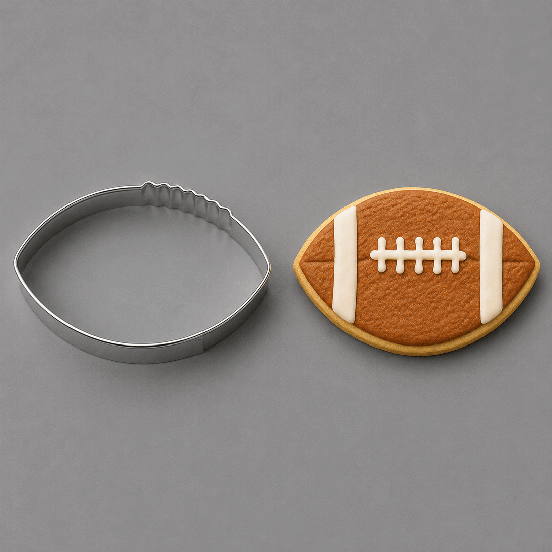 A football-shaped metal cookie cutter shown next to a decorated football-shaped sugar cookie on a smooth gray background from a 3.5 inch Football Metal Cookie Cutter
