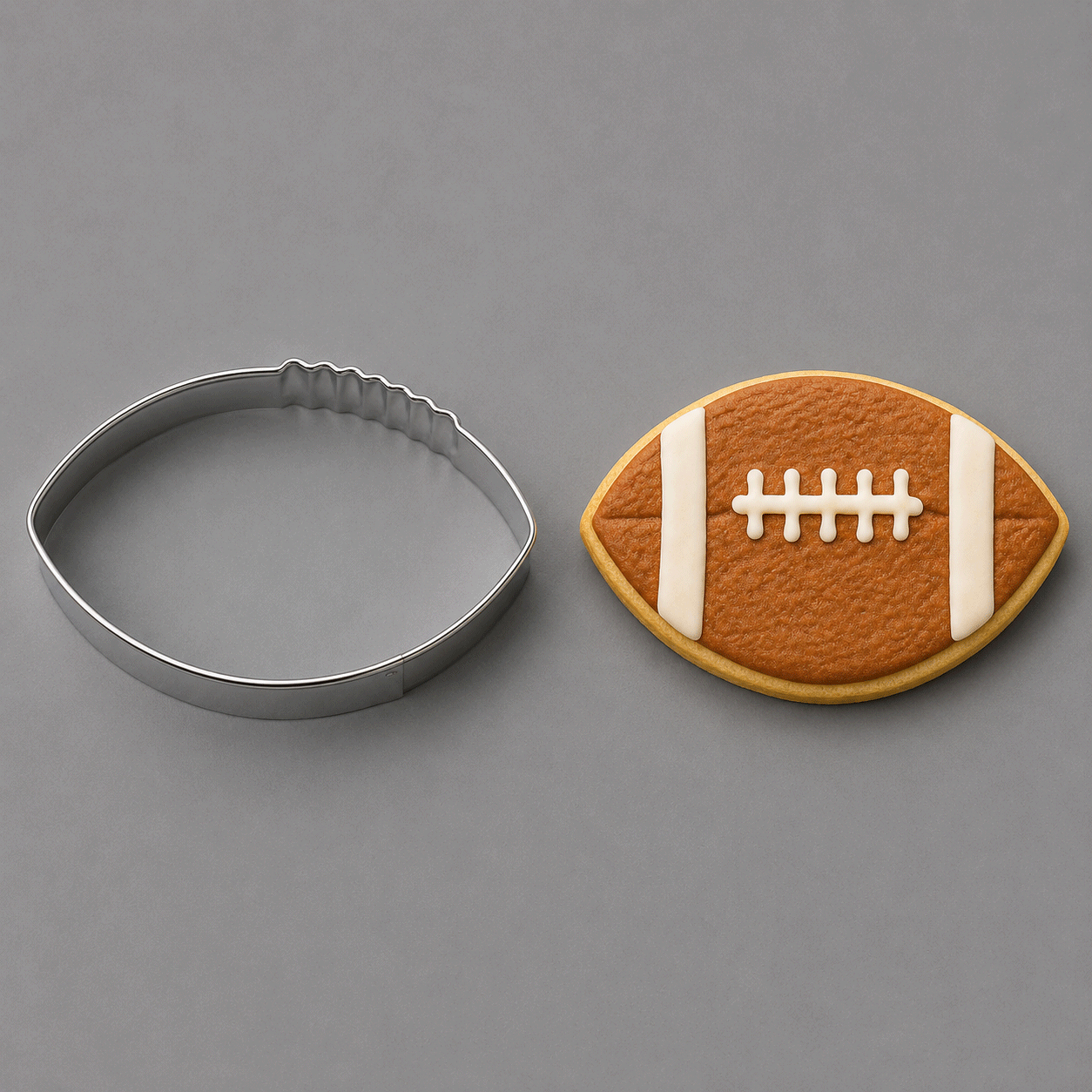 A football-shaped metal cookie cutter shown next to a decorated football-shaped sugar cookie on a smooth gray background from a 3.5 inch Football Metal Cookie Cutter