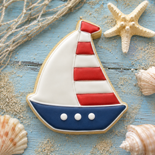 A sugar cookie shaped like a sailboat with white, red, and blue icing, styled on a blue wooden surface with seashells, sand, and rope from a 4 inch Sailboat Metal Cookie Cutter