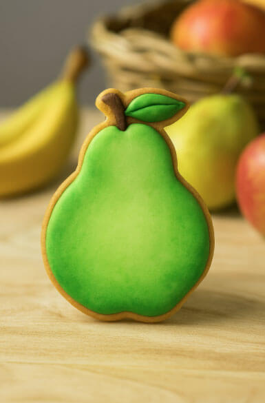 Pear-shaped cookie with green icing and brown stem on a wooden surface with fruits in the background from a 3.75 inch Pear Metal Cookie Cutter