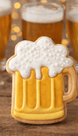 Cookie shaped like a beer mug with foam on a wooden surface with blurred glasses of beer in the background from a 4 inch Root Beer Mug Metal Cookie Cutter