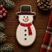 A snowman-shaped decorated cookie on a dark wooden surface, surrounded by festive holiday props including pinecones, evergreen branches, red berries, and a red plaid fabric strip.