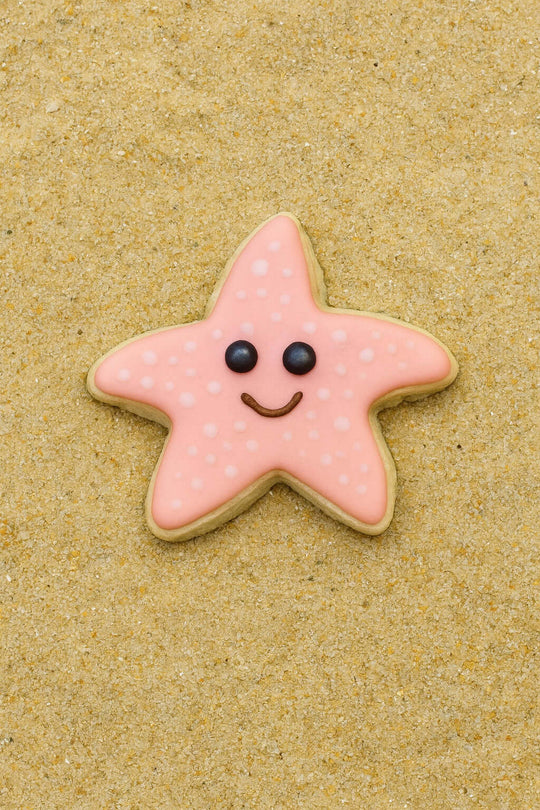 Star-shaped cookie with pink icing and black eyes and mouth on a sandy background