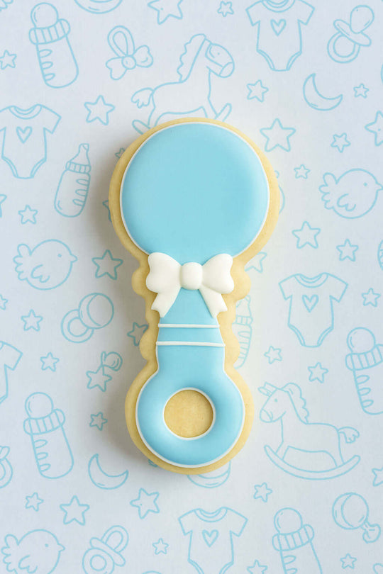 Cookie shaped like a baby rattle with a bow on a light blue background with baby-themed illustrations from a 2 inch Mini Baby Rattle Metal Cookie Cutter