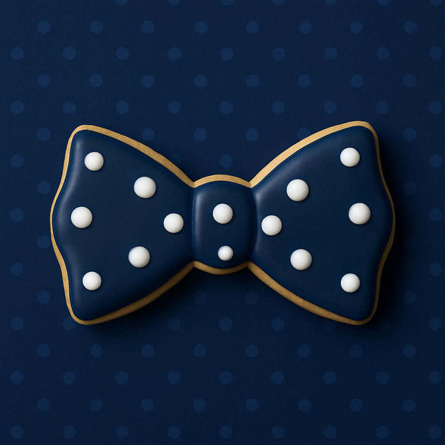 Navy blue bow tie cookie with white dots on a dark blue polka dot background from a 1.75 inch Mini Bow Tie Metal Cookie Cutter