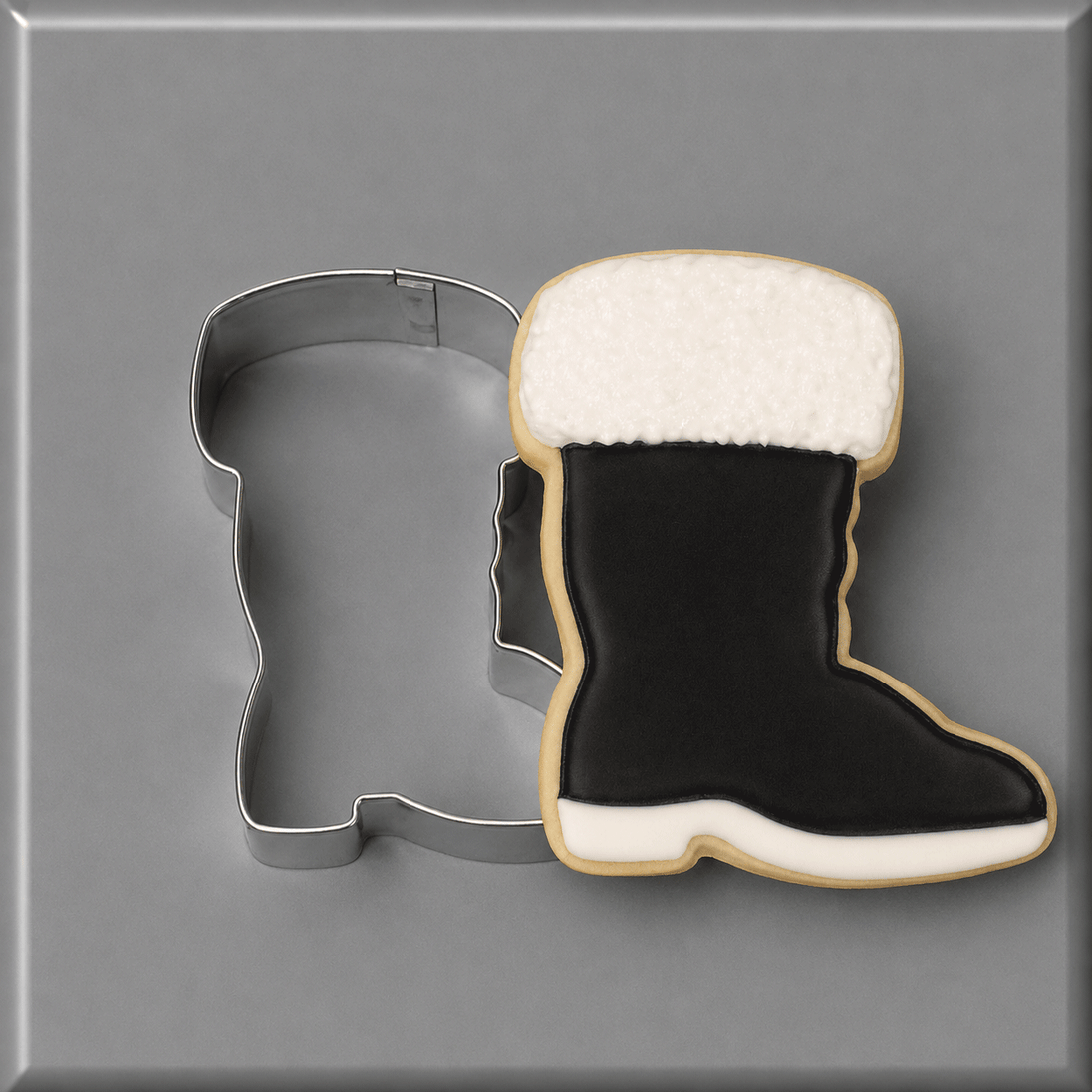 A boot-shaped sugar cookie with black icing, a white faux-fur top, and a white sole, shown next to a 4 inch Santa or Fireman Boot Metal Cookie Cutter on a gray background.