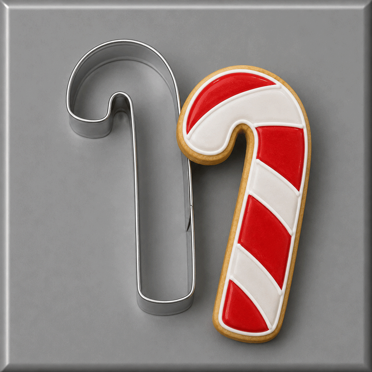 A 5 inch Candy Cane Metal Cookie Cutter shown next to a decorated sugar cookie shaped like a candy cane, on a smooth gray background.