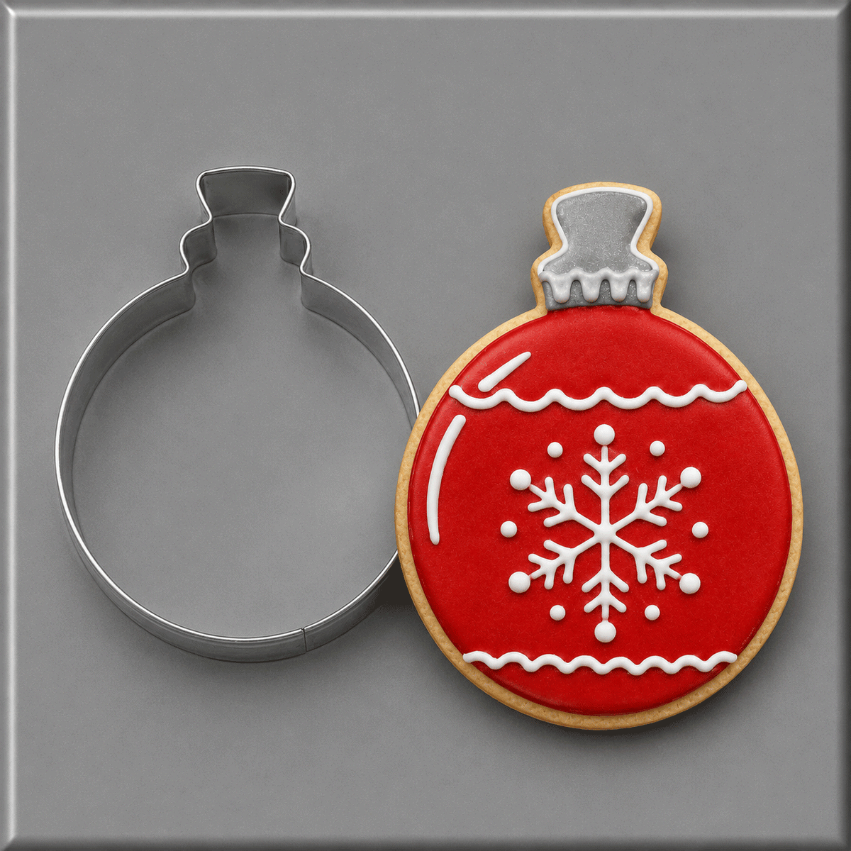 A 4 inch Christmas Ornament Metal Cookie Cutter shown next to a decorated Christmas ornament-shaped sugar cookie with red icing, white icing details, and a gold-toned edge, on a smooth gray background.