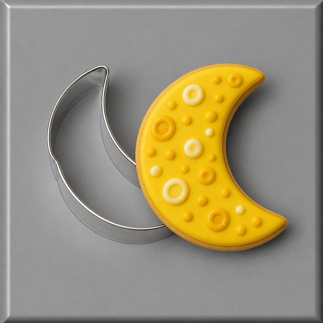 A crescent moon-shaped yellow decorated cookie or fondant piece shown next to a 2.25 inch Mini Crescent Moon Metal Cookie Cutter on a gray background.