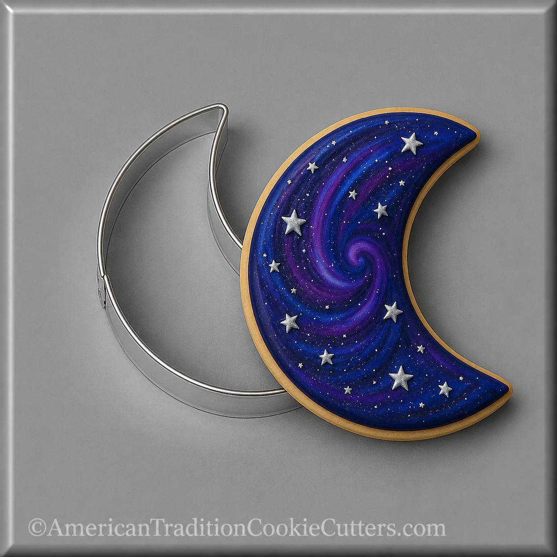 A crescent moon-shaped decorated cookie with a galaxy design in shades of blue, purple, and white, shown next to a 3 inch Crescent Moon Metal Cookie Cutter on a gray background.