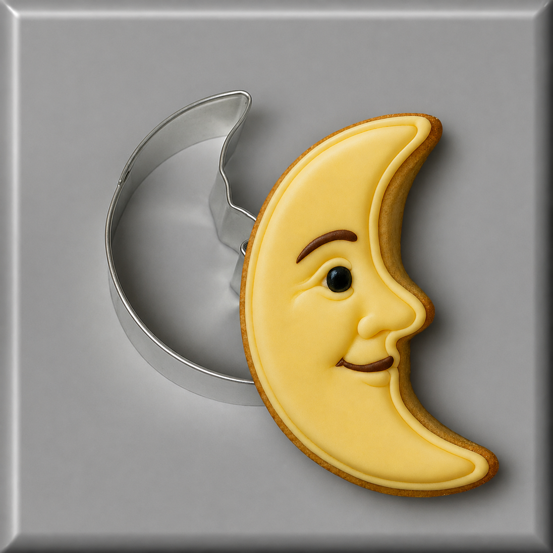 A crescent moon-shaped sugar cookie with a smiling face, decorated in pale yellow icing with brown details, shown next to a3 inch Man in the Moon Metal Cookie Cutter on a gray background.