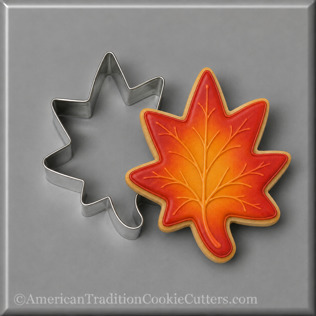 A 3 inch Maple Leaf Metal Cookie Cuttershown beside a decorated maple leaf cookie with orange, red, and yellow icing on a gray background. A watermark at the bottom reads AmericanTraditionCookieCutters.