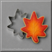 A 3 inch Maple Leaf Metal Cookie Cuttershown beside a decorated maple leaf cookie with orange, red, and yellow icing on a gray background. A watermark at the bottom reads AmericanTraditionCookieCutters.