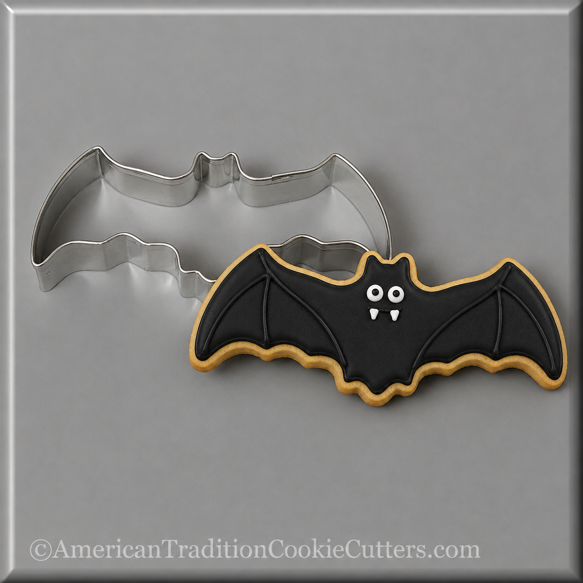 A 4.5 inch Flying Bat Metal Cookie Cutter shown with a decorated bat-shaped cookie underneath on a gray background. The cookie is black with white eyes, white fangs, and a gold outline.