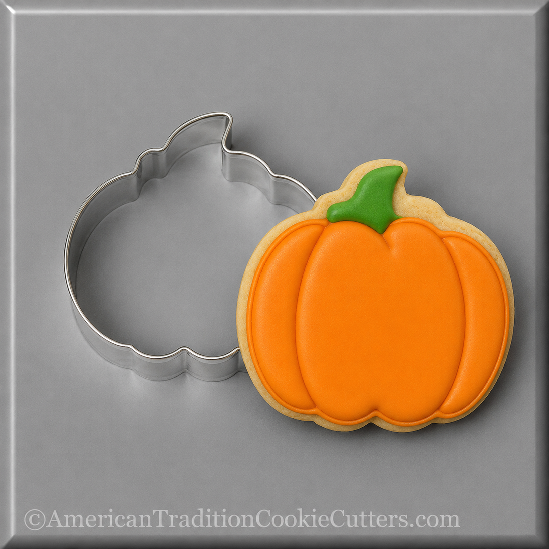A 3 inch Pumpkin Metal Cookie Cutter shown beside a decorated pumpkin-shaped sugar cookie with orange icing and a green stem on a gray background.
