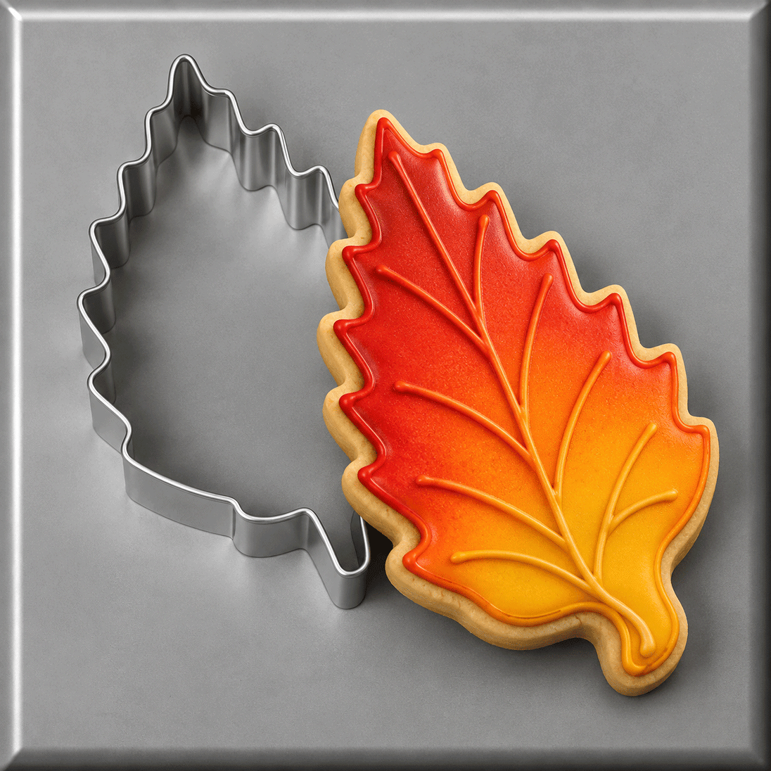 An autumn leaf-shaped sugar cookie with red, orange, and yellow gradient icing sits next to a matching 3.25 inch Aspen Leaf Metal Cookie Cutter on a gray background.