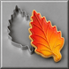 An autumn leaf-shaped sugar cookie with red, orange, and yellow gradient icing sits next to a matching 3.25 inch Aspen Leaf Metal Cookie Cutter on a gray background.