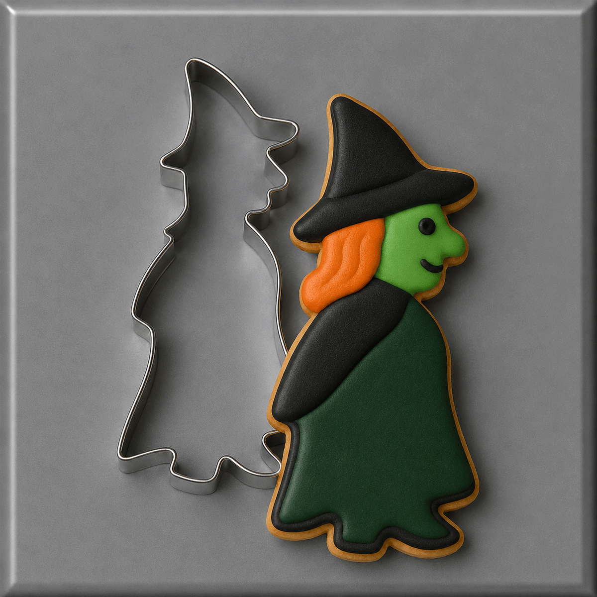A 5 inch Standing Witch Metal Cookie Cutter placed beside a decorated example cookie of a witch with a black hat, green face, orange hair, and dark green dress on a gray background.