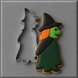 A 5 inch Standing Witch Metal Cookie Cutter placed beside a decorated example cookie of a witch with a black hat, green face, orange hair, and dark green dress on a gray background.