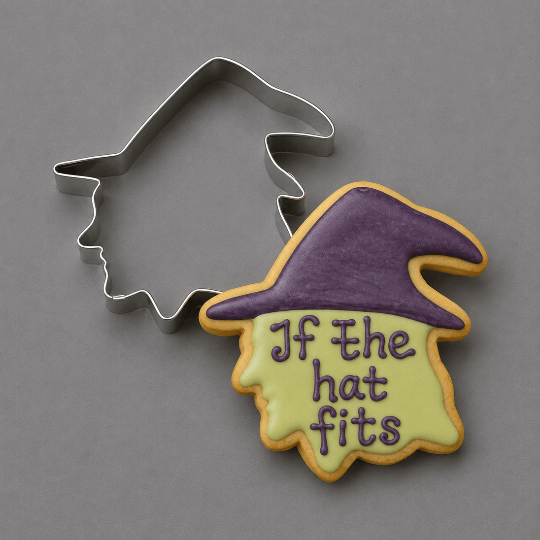 A 4 inch Witch Head Metal Cookie Cutter is shown beside a decorated sugar cookie in the shape of a witch hat with the phrase 'If the hat fits' in purple icing, on a smooth gray background.