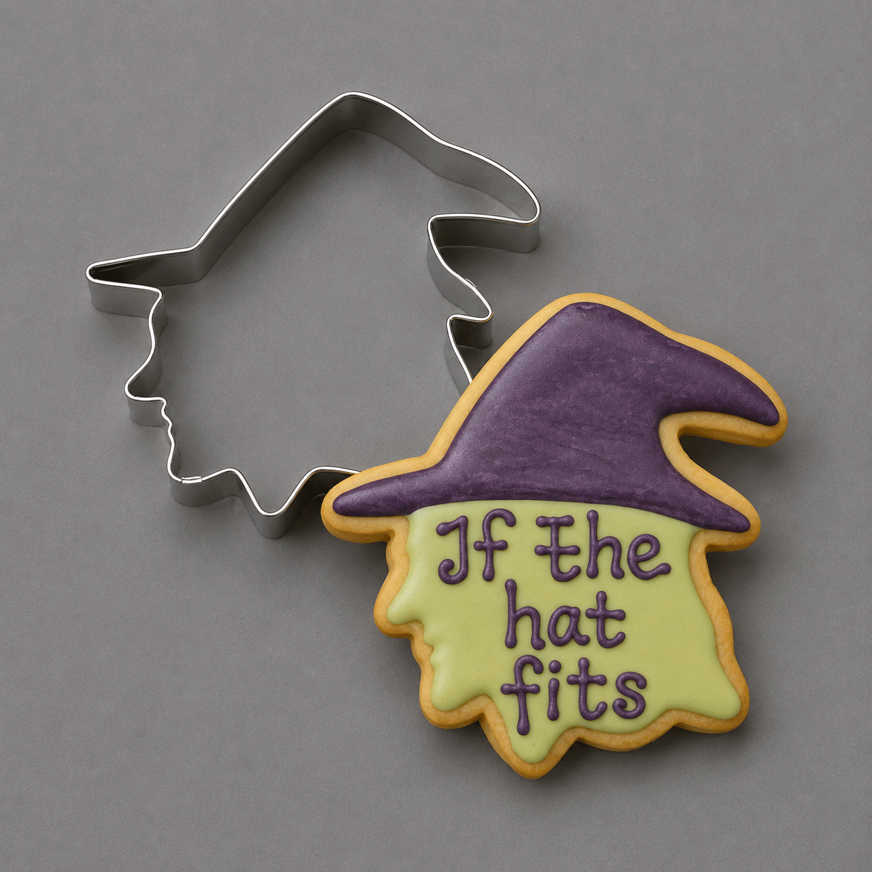 A 4 inch Witch Head Metal Cookie Cutter is shown beside a decorated sugar cookie in the shape of a witch hat with the phrase 'If the hat fits' in purple icing, on a smooth gray background.