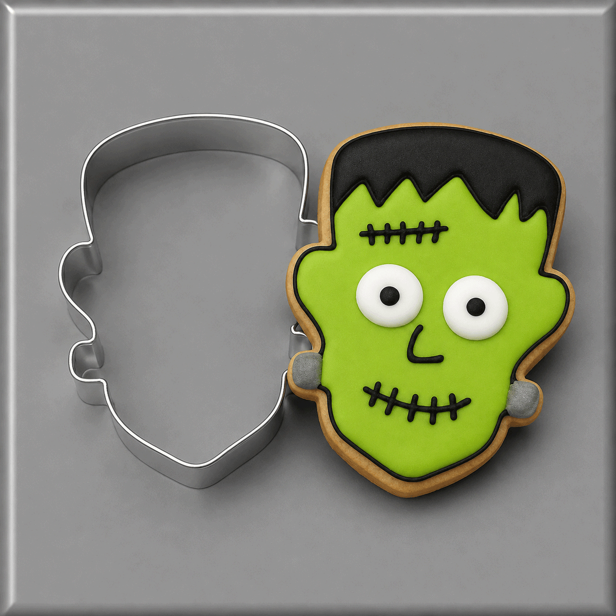 A Frankenstein-themed sugar cookie next to a 3.25 inch Frankenstein Monster Metal Cookie Cutter, set against a neutral gray background.