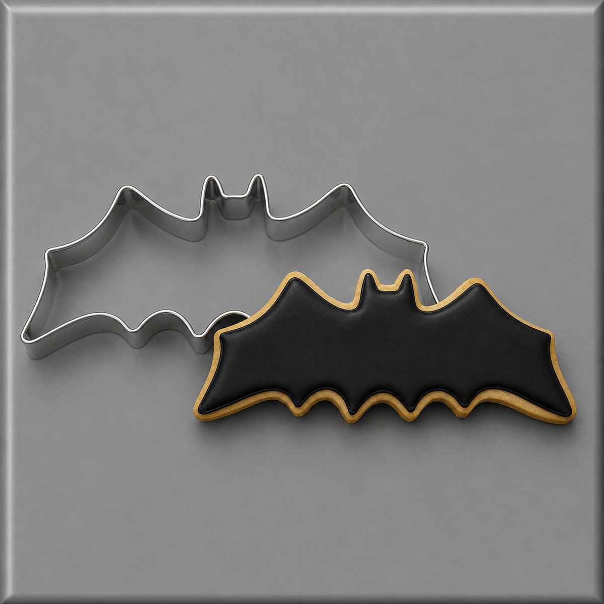 A 4 inch Flying Bat Metal Cookie Cutter in silver metal shown next to a decorated bat-shaped cookie with black icing and a gold border, on a gray background.