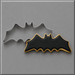 A 4 inch Flying Bat Metal Cookie Cutter in silver metal shown next to a decorated bat-shaped cookie with black icing and a gold border, on a gray background.