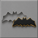A 4 inch Flying Bat Metal Cookie Cutter in silver metal shown next to a decorated bat-shaped cookie with black icing and a gold border, on a gray background.