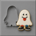 A ghost-costume-shaped sugar cookie with white icing, black eyes and mouth, a red tongue sticking out, and black-and-blue shoes, shown next to a 4 inch Ghost Halloween Costume Metal Cookie Cutter on a gray background.
