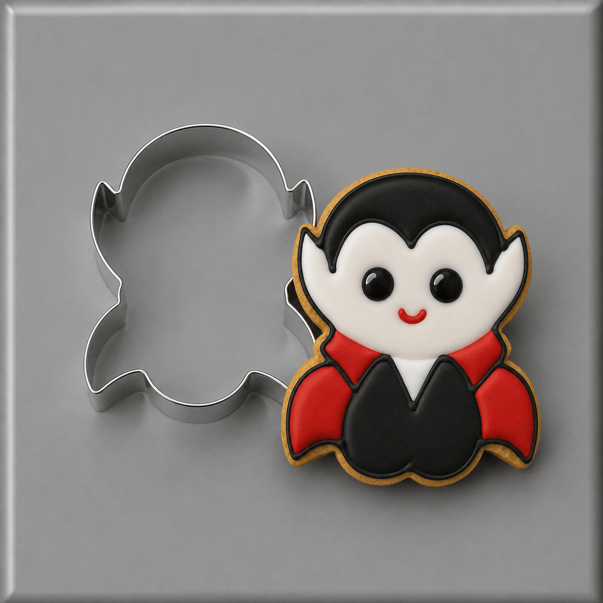 A 4 inch Count Dracula Vampire Metal Cookie Cutter shown next to a decorated vampire-shaped cookie on a smooth gray surface.