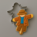 A decorated scarecrow-shaped cookie or cookie cutter design shown on a plain gray background. The figure wears a brown hat, brown jacket and pants, a bright blue shirt, and yellow hands, with a 4 inch Scarecrow Metal Cookie Cutter outline behind it.