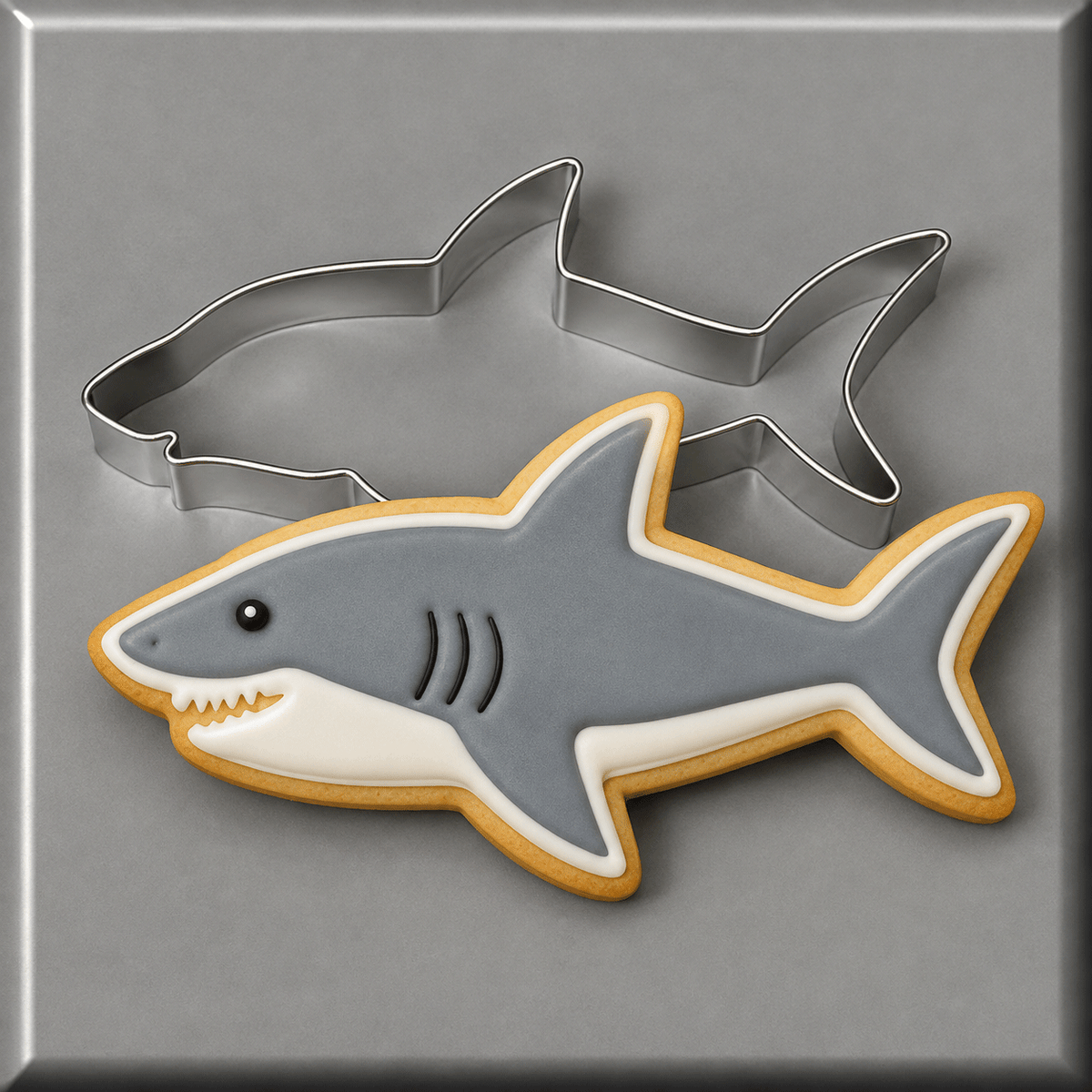A shark-shaped sugar cookie with gray, white, and tan icing, shown with a 4 inch Shark Metal Cookie Cutter on a gray background.