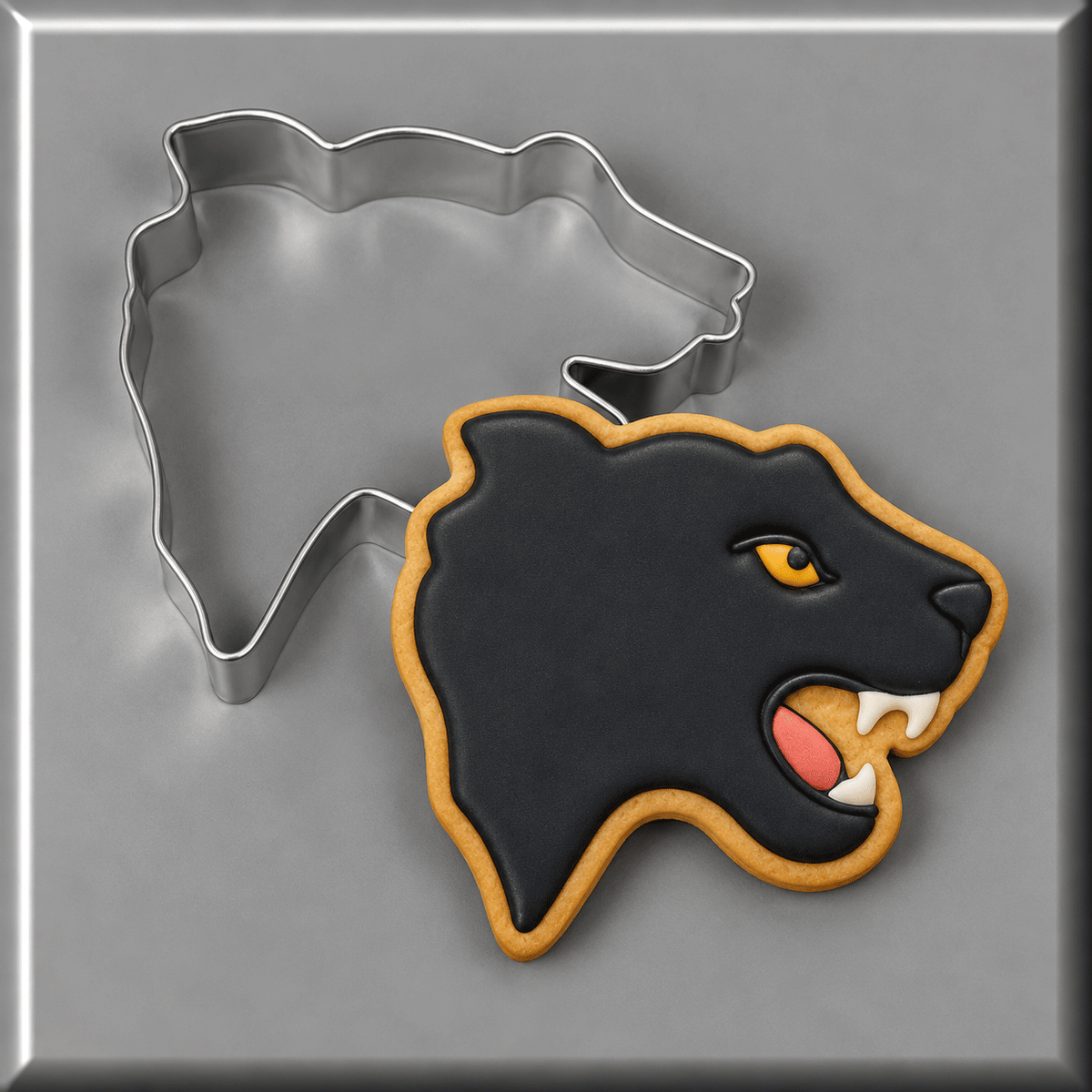 A black panther cookie with gold icing and a pink tongue, shown next to a 3.5 inch Panther Head Metal Cookie Cutter on a gray background.