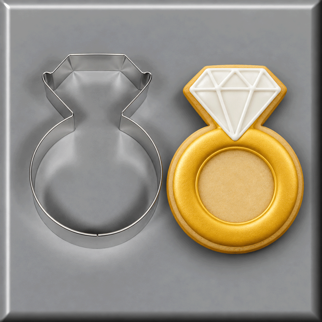 A 3.75 inch Diamond Engagement Ring Metal Cookie Cutter shown next to a decorated ring-shaped sugar cookie with a white diamond design and gold band, on a gray background.