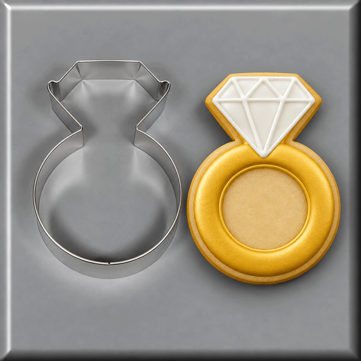 A 3.75 inch Diamond Engagement Ring Metal Cookie Cutter shown next to a decorated ring-shaped sugar cookie with a white diamond design and gold band, on a gray background.